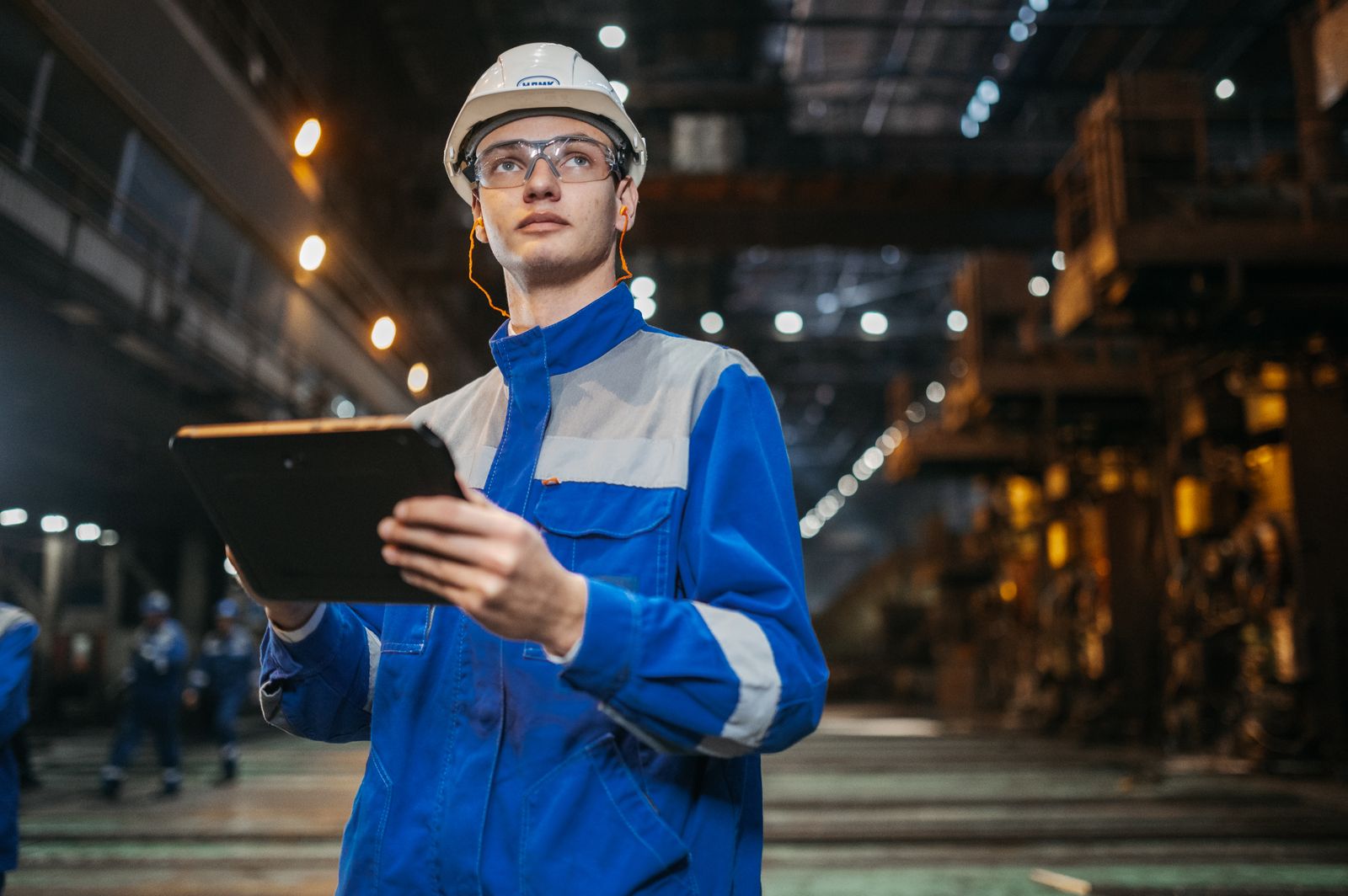 Engineer in safety gear using a tablet inside an industrial factory.